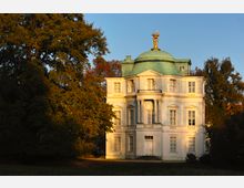 Historisches Gebäude mit weißer Fassade, einer Kuppel aus grünem Kupfer und einer goldenen Statue an der Spitze, umgeben von großen Bäumen in einem parkähnlichen Gelände bei Sonnenuntergang.