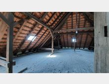 Dachbodenansicht mit sichtbarer Holzkonstruktion und roten Ziegeln unter dem Dach. Der Boden besteht aus alten Holzdielen, und ein kleines Dachfenster lässt Tageslicht herein.