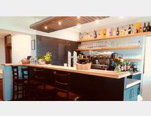 Eine moderne Bar mit einer hölzernen Theke, schwarzen Barhockern und Regalen, die Gläser und Flaschen enthalten. Im Hintergrund stehen eine Kaffeemaschine und Zapfanlagen, vorne sind Dekorationen wie Pflanzen und ein Blumenstrauß.