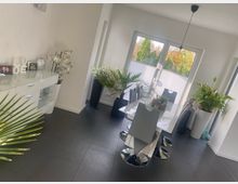 Heller, moderner Essbereich mit dunklen Fliesen, einem weißen Esstisch mit Stühlen und einer schwarzen Hängelampe. Große Fenster mit teilweiser Sichtschutzfolie öffnen den Blick ins Grün; mehrere große Topfpflanzen und ein weißes Sideboard mit Deko stehen an den Wänden.