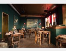 Gemütlicher Innenraum eines Cafés mit dunkelgrünen Wänden, Holzböden und Tischen aus Holz, die von schlichten Stühlen umgeben sind. Kerzen und Blumen auf den Tischen sowie eine große Wandmalerei und rote Vorhänge prägen die Atmosphäre, während die Beleuchtung dezent ist.