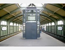 Bahnhof mit überdachter Bahnsteighalle und gewölbtem Dach aus Stahlträgern, im Zentrum ein gläserner Fahrstuhl. Zwei parallel verlaufende Gleise und Stationsschilder mit der Aufschrift „Schönhauser Allee“ sind sichtbar.