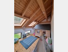 Holzvertäfeltes Kinderzimmer mit schräger Holzdecke und sichtbaren Balken. Das Zimmer enthält ein Spielhäuschen aus Holz, ein graues Tipi und eine blaue Spielmatte; große Fenster und Dachfenster lassen viel Tageslicht herein.