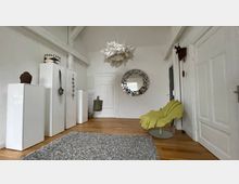 Helle Wohnzimmerecke mit Holzfußboden, einer modernen weißen Hängelampe, einem großen runden Spiegel mit dekorativem Rahmen an der Wand und einem grünen Designersessel. Links befinden sich weiße Schränke mit Deko-Objekten, rechts eine Zimmertür und an einer weiteren Tür hängt ein Wandanhänger.