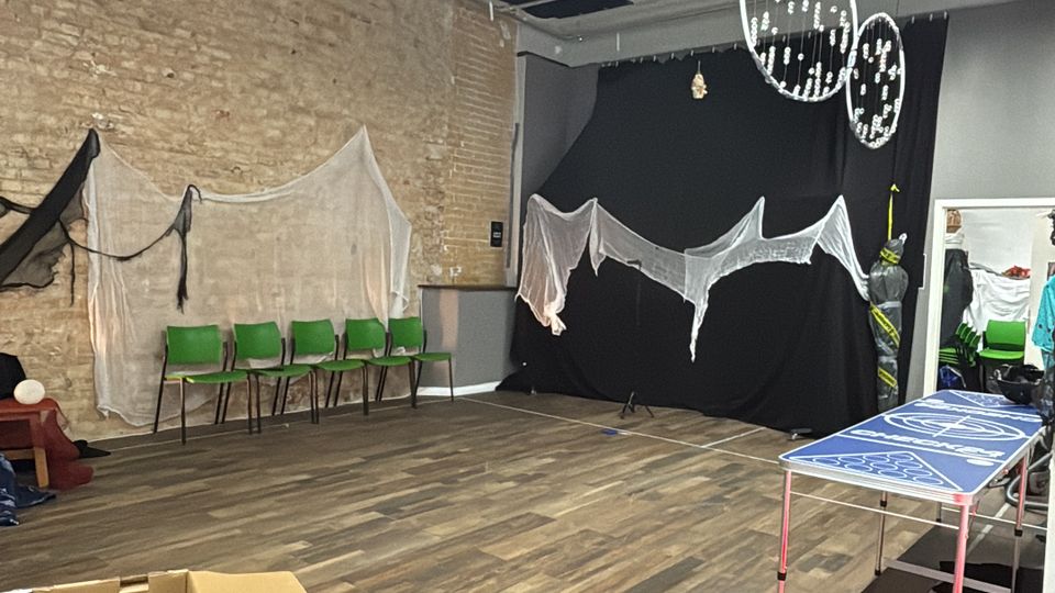Spacious indoor studio or event room with wood-look flooring, an exposed brick wall, and a grid ceiling with fluorescent strip lights and hanging circular decorations. Green chairs line one wall, sheer fabric drapes hang on the walls, and boxes and equipment are scattered around the open floor.