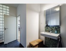 Kleines Badezimmer mit einem rechteckigen Waschbecken, einem beleuchteten Spiegelschrank und einer Wand mit grüner Mosaikfliesenverkleidung. Links ist eine offene Tür, die in einen Raum mit Holzboden führt, und daneben eine Wand aus Glasbausteinen.