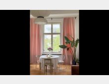 Heller Wohnraum mit einem großen Fenster, flankiert von rosafarbenen Vorhängen. Im Vordergrund steht ein runder Tisch mit weißer Tischdecke, umgeben von Metallstühlen, daneben große grüne Zimmerpflanzen und eine papierartige Hängeleuchte.