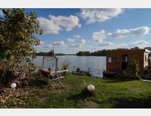 Grünfläche am Ufer eines Sees mit einer Schaukel, einem kleinen Holzsteg und einem hölzernen Hausboot auf der rechten Seite. Im Hintergrund sind ruhiges Wasser, bewaldete Inseln und ein blauer Himmel mit weißen Wolken zu sehen.
