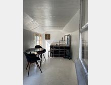 Kleiner, schmaler Raum mit einem weißen Fußboden und schrägen Wänden. Im Vordergrund steht ein runder Tisch mit zwei schwarzen Stühlen, im Hintergrund ein Bett mit buntem Bettzeug und einem Regal darunter. Ein Fenster auf der linken Seite lässt Tageslicht herein, und ein Ventilator steht rechts neben dem Bett.