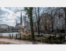 Städtischer Park mit kahlen Bäumen und einem Zaun im Vordergrund, dahinter ein kleiner Fluss oder Teich. Im Hintergrund ist eine neugotische Backsteinkirche mit hohem Turm sowie Wohngebäude sichtbar.