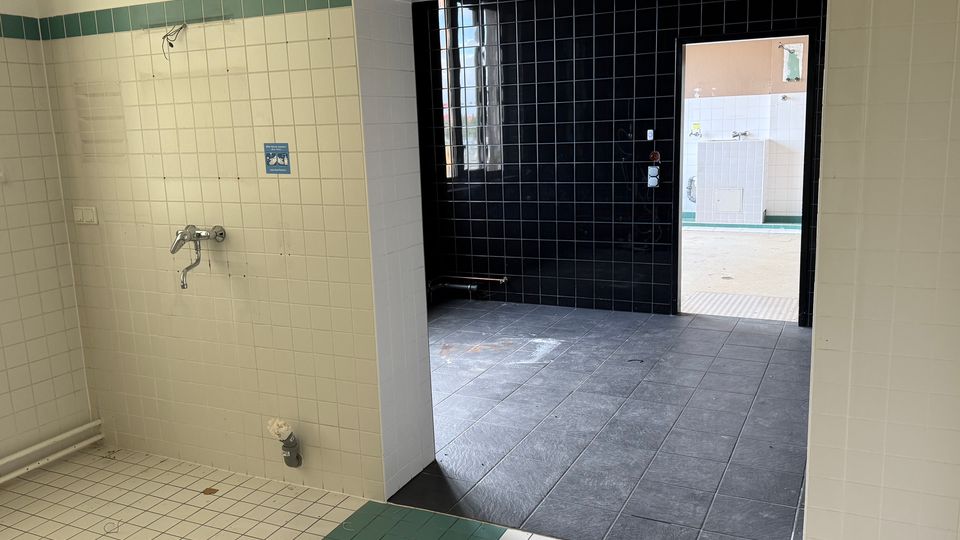 Gefliester Innenraum mit weißen Wandfliesen und grünem Rand, links mit einem Wandwasserhahn und Leitungen. Im Durchgang liegt ein dunkler, quadratisch gefliester Bereich mit schwarzer Rückwand und Fenster, der zu einem weiteren hell gefliesten Nebenraum führt.