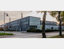 Industriegebäude mit grauer Fassade und großen Fensterreihen, flankiert von zwei Backsteinschornsteinen. Im Vordergrund befinden sich eine Straßenkreuzung, Straßenlaternen und Bäume.