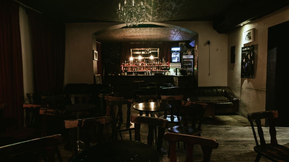 Dimly lit bar interior with worn wooden floors, scattered small round tables and chairs, and a leather bench along the wall. An arched ceiling with a chandelier frames a back bar lined with bottles, hanging lights, and a glass-front drink refrigerator.