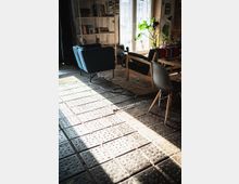 Ein Wohnzimmer mit modernen Möbeln, darunter ein blauer Sessel, ein Holzbett mit schwarzem Polster und ein grauer Kunststoffstuhl. Im Hintergrund sind ein Bücherregal, eine großzügige Fensterfront mit Vorhängen und Zimmerpflanzen zu sehen; der Boden besteht aus gemusterten Fliesen und einem großen Teppich.