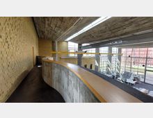 Innenansicht eines modernen Gebäudes mit gelben Ziegelwänden, einer geschwungenen Betonbrüstung mit Holzabdeckung und einer hohen Decke aus Sichtbeton. Große Glasfenster lassen viel Tageslicht herein und bieten Blick auf einen grünen Außenbereich.