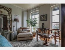 Wohnzimmer mit klassischem Interieur: Im Mittelpunkt stehen ein beigefarbener Sessel mit Hocker, ein kleiner Holztisch mit Schachbrett und zwei Stühle mit Korbgeflecht. An der Wand hängen Bilder, rechts und links befinden sich große Fenster mit Blick auf einen Balkon und die Berliner Fernsehturm-Silhouette. Ein großer Zierspiegel und grüne Zimmerpflanzen ergänzen den Raum.