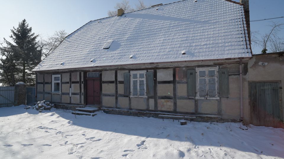 Vorderansicht eines eingeschossigen Fachwerkhauses mit großem, schneebedecktem Satteldach und kleinen Fenstern mit Fensterläden. Davor liegt ein verschneiter Hof, im Hintergrund stehen kahle Bäume und ein Zaun neben dem Gebäude.