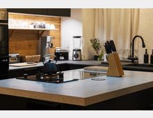 Moderne Küche mit einer Arbeitsinsel aus hellem Stein, darauf ein Gaskochfeld mit einer schwarzen Teekanne und ein Messerblock aus Holz. Im Hintergrund schwarze Schränke, Holzregale mit Geschirr, eine schwarze Armatur, ein Toaster und eine Küchenmaschine.