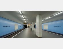 U-Bahn-Station mit blauen Kachelwänden, rechteckigen Säulen mit Mosaikfliesen und zwei Gleisen. Im Hintergrund befinden sich eine Uhr, ein Anzeigeschild und eine Treppe, die zu einem Ausgang führt.
