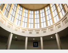 Runder Innenraum mit hoher Decke und großen Glasfenstern in Bogenform, die Tageslicht einlassen. Im unteren Bereich sind weiße Wände mit Lüftungsgittern zu sehen, darüber hängt ein Schild mit der Zahl "13".