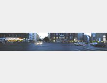 Weitläufige Straßenkreuzung in einer urbanen Umgebung, umgeben von mehrstöckigen Gebäuden mit Geschäften und Restaurants im Erdgeschoss, darunter leuchtende Reklametafeln. Mehrere Autos, Fahrräder und Fußgänger sind auf der Straße und an den Gehwegen zu sehen, während Straßenlaternen und die Dämmerung die Szene beleuchten.