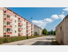 Mehrstöckige Wohnhäuser mit hellen Fassaden und roten Balkonen stehen entlang einer ruhigen Straße mit Gehwegen und niedrigen Büschen. Im Hintergrund sind Bäume und einzelne Gebäude unter blauem Himmel mit weißen Wolken zu sehen.