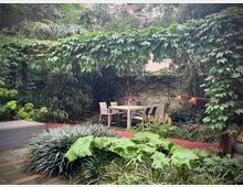 Ein kleiner, begrünter Innenhof mit üppigen Pflanzen, Kletterpflanzen an einer Pergola und großblättrigen Gewächsen. In der Mitte steht ein weißer Gartentisch mit Stühlen, umgeben von Pflastersteinen und einer Holzterrasse.