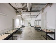 Ein schlichter, weiß gestrichener Raum mit leerstehenden Glasmontrevitrinen entlang beider Wände. An der Decke befinden sich freiliegende Rohre und Lüftungsleitungen, während der Boden aus abgenutztem Beton besteht.