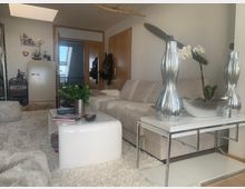 Wohnzimmer mit modernem Design: ein beiges Sofa vor hellen Holztüren, ein weißer Couchtisch auf einem flauschigen Teppich, eine silberne Dekovase auf einem Beistelltisch und ein Wandpropeller als dekoratives Element, mit natürlichem Licht durch ein Dachfenster. Im Hintergrund stehen eine Retro-Kühltruhe und ein kleiner Barwagen.