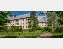 Mehrfamilienhäuser mit hellem Putz und grauem Satteldach, umgeben von Bäumen, Sträuchern und einer Wiese im Vordergrund; ein geteilter Gehweg führt durch die grüne Umgebung.