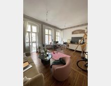 Ein Wohnzimmer in einer Altbauwohnung mit hohen Decken, verzierten Stuckleisten und großen, bogenförmigen Fenstern. Der Raum ist mit Holzdielen, einer Sitzecke, einem runden Couchtisch, einem Teppich sowie einer Leiter zur kleinen Regalecke ausgestattet; im Hintergrund stehen eine Kommode, ein Fernseher und Dekorationen.