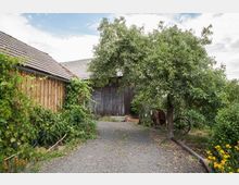 Hof mit einem Schotterweg, gesäumt von einem Baum und blühenden Pflanzen. Im Hintergrund befinden sich eine Holzwand einer Scheune und ein Holzschuppen mit Ziegeldach.