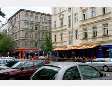 Straßenkreuzung mit Altbauten und Cafés. Links ein graues, verziertes Gebäude, rechts ein helles Gebäude mit orange-weiß gestreiften Markisen, darunter ein Außenbereich mit Tischen und Gästen. Im Vordergrund parkende Autos.