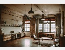 Großzügiger Raum im Industriestil mit freiliegender Backsteinwand, großen Fenstern mit grauen Vorhängen und Holzboden. Die Einrichtung umfasst Holzmöbel, Regale mit Deko-Objekten und Industrielampen an der Decke.