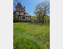 Historisches Gebäude mit Fachwerkelementen und spitzem Turmdach in einer ländlichen Umgebung, umgeben von einer Wiese und mehreren großen Bäumen unter klarem blauem Himmel.