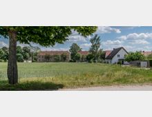 Ländliche Siedlung mit einem Baum im Vordergrund, einem Feld in der Mitte und Häusern mit roten Dächern im Hintergrund. Die Umgebung ist von Bäumen und Grünflächen umgeben, der Himmel ist leicht bewölkt.
