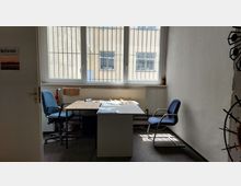 Ein kleiner, einfacher Büroraum mit zwei blauen Stühlen und einem Schreibtisch, auf dem Papiere und Unterlagen liegen. Im Hintergrund befindet sich ein großes, vergittertes Fenster, durch das ein Blick auf ein gegenüberliegendes Gebäude möglich ist.