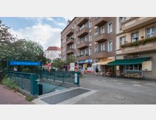 Eingang zur U-Bahn-Station "Amrumer Straße" mit einem blauen Schild, umgeben von mehrstöckigen Wohn- und Geschäftsgebäuden mit Geschäften und Cafés im Erdgeschoss. Vor der Station befinden sich Bäume und ein kleiner Platz mit Sitzmöglichkeiten.