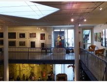 Innenansicht eines zweistöckigen Museums mit Oberlicht. Die obere Ebene zeigt Gemälde an den Wänden und eine Figurenausstellung in der Mitte, während die untere Ebene Vitrinen mit Figuren und Exponaten enthält.