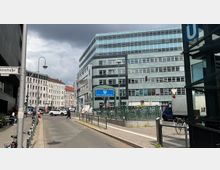 Straßenszene am Rosa-Luxemburg-Platz mit modernen und historischen Gebäuden, einem blauen U-Bahn-Schild und einer Ampel. Im Vordergrund Gehwege, geparkte Fahrräder und eine U-Bahn-Zugangstreppe.