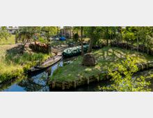 Kleiner Wasserkanal in einem ländlichen Garten mit einem flachen Holzboot und einem weiteren Boot, das mit einer Plane abgedeckt ist. Umgeben von Bäumen, Holzzäunen und einer Grasfläche, auf der ein Heuhaufen liegt.