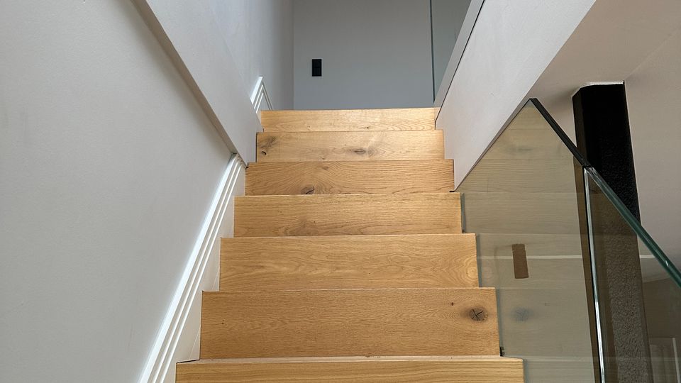 Innenaufnahme eines modernen Treppenhauses mit hellen Holzstufen, weißen Wänden und einem Glasgeländer. Die Treppe führt nach oben zu einem offenen Flur mit Glasbrüstung und sichtbarem dunklem Deckenbalken.