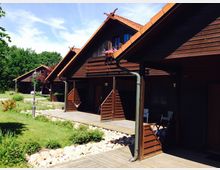 Holzhäuser mit roten Ziegeldächern in einer gepflegten, grünen Gartenanlage. Vor den Gebäuden befinden sich Holzbalkone, ein gepflasterter Weg und dekorative Blumenbeete.