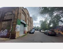 Ehemaliges Industriegebäude aus Backstein mit hohen, bogenförmigen Fenstern, dessen Wände teils mit Graffiti versehen sind. Vor dem Gebäude befinden sich mehrere parkende Autos, und seitlich ist eine bemalte Mauer mit urbaner Kunst sowie einige Bäume zu sehen.