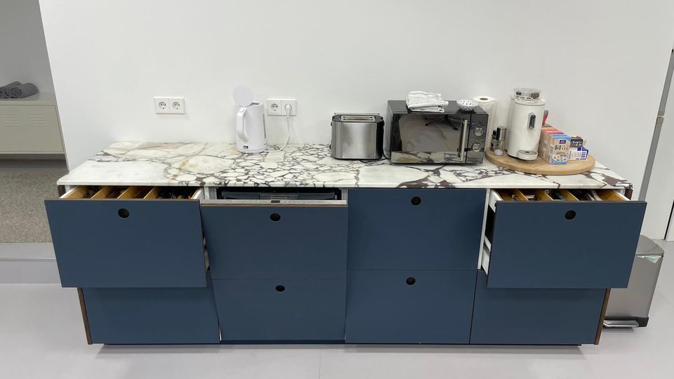 Kitchenette counter with a white marble-look surface and blue lower cabinets, several drawers open to show utensil dividers. Small appliances including a kettle, toaster, microwave, and coffee machine sit along the backsplash beneath wall outlets.
