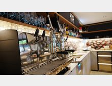 Innenansicht einer modernen Café- oder Bar-Theke mit großer Edelstahl-Espressomaschine im Vordergrund, darüber Regale mit Gläsern und Spirituosenflaschen. Im Hintergrund sind eine beleuchtete Arbeitsfläche, Spüle und eine geflieste Rückwand mit rechteckigem Muster zu sehen.