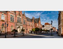 Straßenszene mit einem historischen Gebäude aus rotem Backstein mit hohen, verzierten Fenstern und einem Haupteingang mit steinernen Details auf der linken Seite. Rechts befinden sich weitere Gebäude, darunter ein helles Wohnhaus, mit Bäumen und Straßenlaternen entlang der gepflasterten Straße.