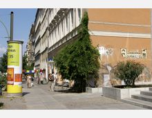 Straßenszene in einer urbanen Umgebung mit einem gelben Werbesäulenplakat im Vordergrund, einer Reihe historischer Stadthäuser auf der linken Seite und einem begrünten Gehweg. Rechts steht eine mit Graffiti versehene Wand, ein Baum und einige Sitzgelegenheiten, während im Hintergrund Menschen entlang der Straße gehen.