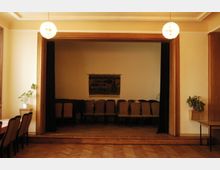 Ein kleiner Veranstaltungsraum mit einer erhöhten Bühne im Hintergrund, auf der mehrere Stühle hintereinander aufgestellt sind. Der Raum hat Holzböden, helle Wände, Pflanzen an den Seiten und hängt in der Mitte eine kunstvolle Wanddekoration.