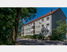 Mehrfamilienhaus mit drei Stockwerken und einem roten Satteldach, umgeben von Bäumen und Grünflächen. An der Vorderseite des Gebäudes befinden sich Balkone, eine Straße mit Gehweg verläuft entlang der Fassade.